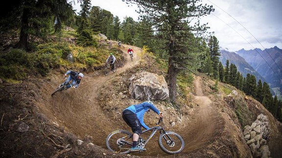 Mountain biker on a winding trail in the forest. In the background, high mountains and green trees can be seen. - CORSO living Aparthotel - Sölden