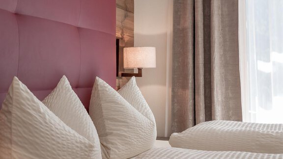 A modern bedroom with a purple headboard and two large pillows. The room has light curtains and a stylish lamp. - The Peak Sölden - Sölden