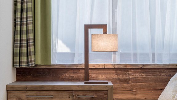 A modern lamp stands on a wooden table next to a window with light curtains. The room has a cozy and inviting atmosphere. - The Peak Sölden - Sölden