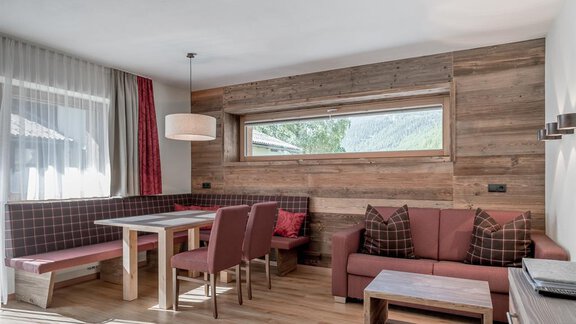 A modern living and dining room with a large window and beautiful wood paneling. There is a dining table, chairs, and a cozy couch. - The Peak Sölden - Sölden