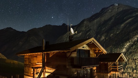 A cozy wooden house under a clear starry sky. The surrounding mountains are dimly lit. - The Peak Sölden - Sölden