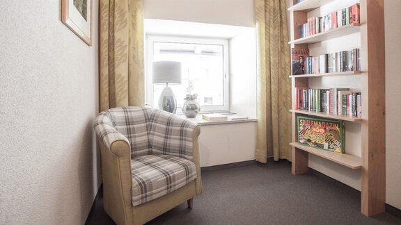 A cozy reading nook with a checked armchair and a bookshelf. The room is bright and inviting with curtains and a lamp. - Garni Ötztal ROOMS & APARTS - Zwieselstein