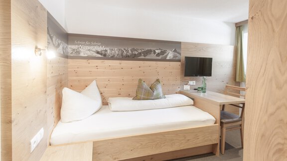 A modern, cozy hotel room with wooden interior. There is a single bed, a table, and a television mounted on the wall. - Garni Ötztal ROOMS & APARTS - Zwieselstein
