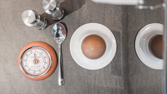 A table with dishes and utensils for breakfast. There is a thermometer, salt and pepper shakers, a spoon, and an egg in a white egg cup. - Garni Ötztal ROOMS & APARTS - Zwieselstein