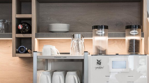 A modern café row with an elegant coffee machine and white cups. In the background, wooden shelves and glasses are visible. - Garni Ötztal ROOMS & APARTS - Zwieselstein