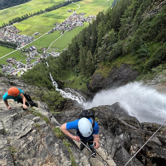 Two climbers are ascending a steep cliff. In the background, one can see a valley with a town and a waterfall. - Alpintreff - Längenfeld