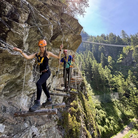 Zwei Personen klettern auf einem schmalen Steg an einer Felswand entlang. Im Hintergrund sind grüne Wälder und ein blauer Himmel zu sehen. - Alpintreff - Längenfeld