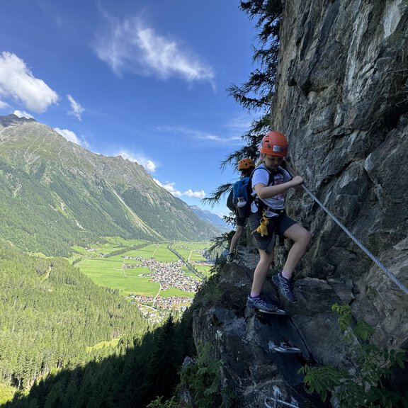 Ein Kind klettert an einer steilen Felswand mit Hilfe eines Seils. Im Hintergrund erstreckt sich eine grüne Tallandschaft unter blauem Himmel. - Alpintreff - Längenfeld