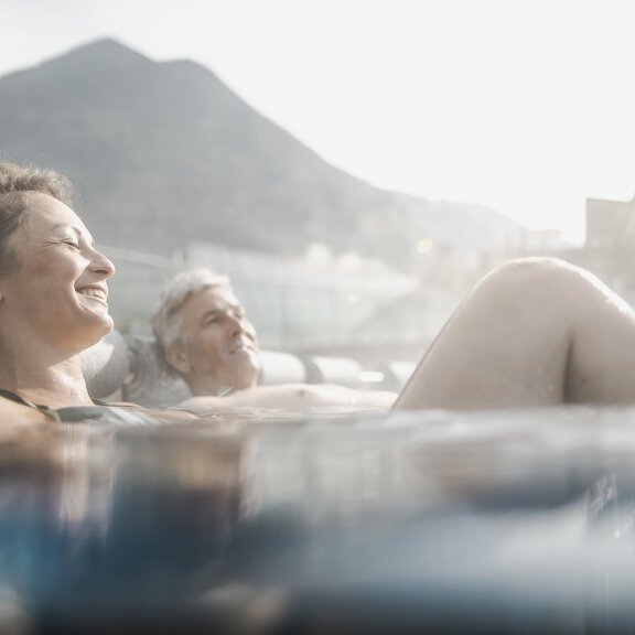 A relaxed couple enjoys the warm water of a hot tub with a view of the mountains. The sun is shining, creating a pleasant atmosphere. - AQUA DOME - Tirol Therme Längenfeld - Längenfeld