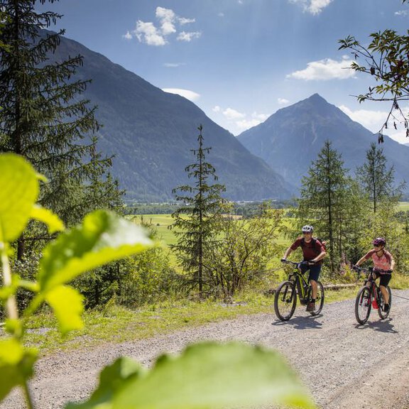 Two cyclists ride along a gravel path in a picturesque mountain landscape. In the background, high mountains and a bright blue sky can be seen. - Lohmann Längenfeld - Längenfeld