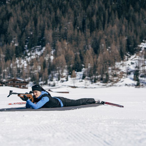 A person is lying in the snow and aiming with a rifle. In the background, trees and buildings are visible in a wintry landscape. - ÖTZTAL TOURISMUS - Info Längenfeld (Alpinzeit) - Längenfeld