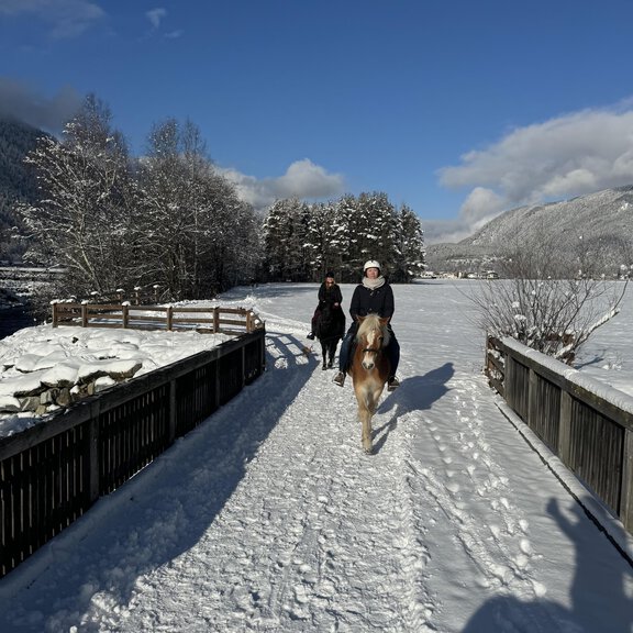 Two people are riding on a snow-covered path. In the background, there are snow-covered trees and a blue sky. - Reitstall Pegasus Längenfeld - Längenfeld