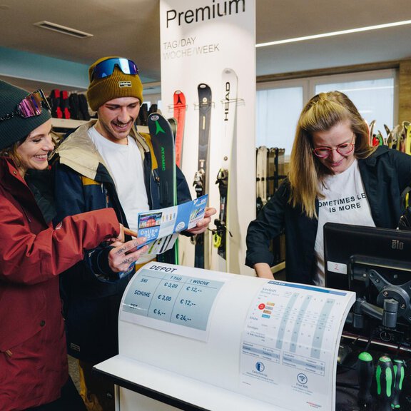 Two customers are looking at skiing equipment in a sporting goods store. An employee is assisting them at the computer. - Sport Glanzer Längenfeld - Längenfeld