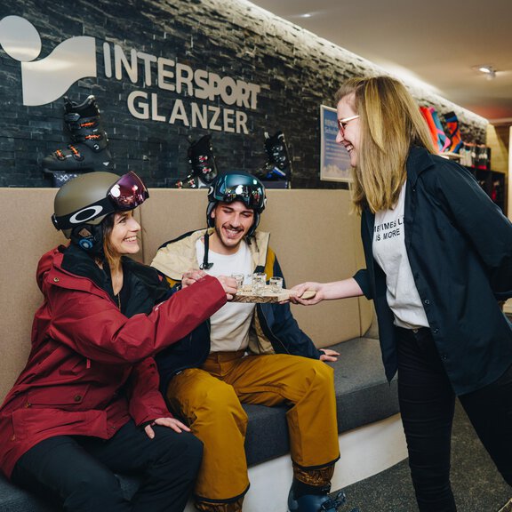 Three people are sitting in a ski service area and smiling. An employee from Intersport Glanzer is handing them something. - Sport Glanzer Längenfeld - Längenfeld