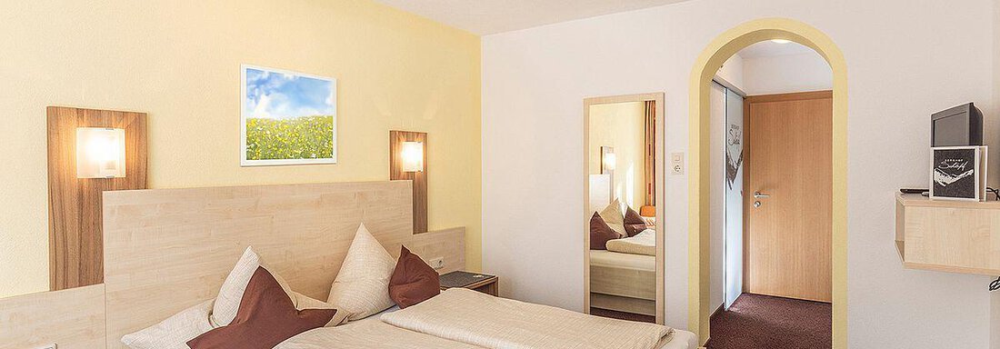 A cozy hotel room with a double bed and a large mirror. The walls are painted in soft colors and a window showcases a beautiful landscape. - Berghof Schöpf - Gries