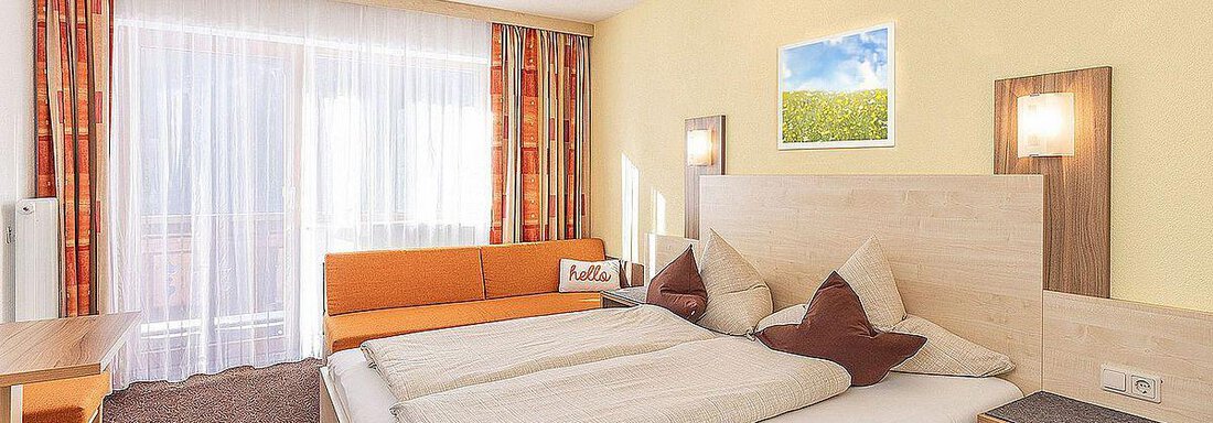 A bright hotel room with a large double bed and a comfortable sofa. The walls are painted in warm colors and large windows provide plenty of daylight. - Berghof Schöpf - Gries