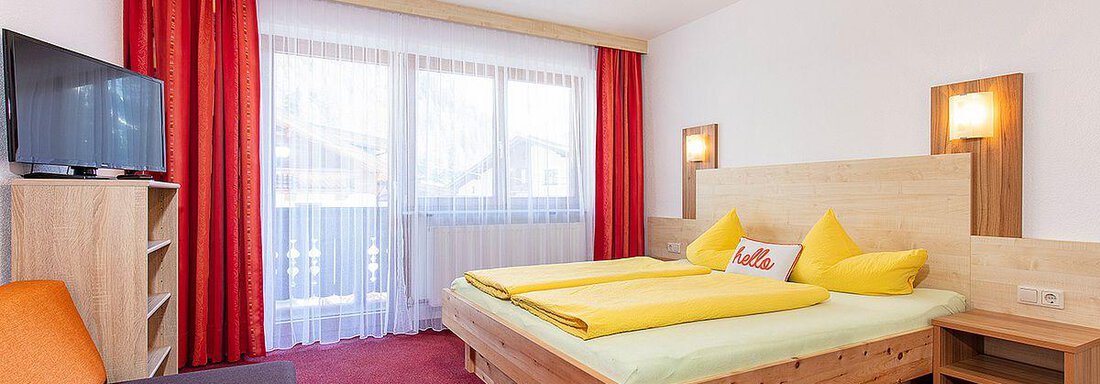 A modern hotel room with a double bed and two yellow pillows. The room has a TV, a balcony, and large windows with curtains. - Berghof Schöpf - Gries