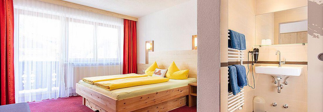 A bright room with a double bed and yellow pillows. In the background, a modern bathroom can be seen. - Berghof Schöpf - Gries