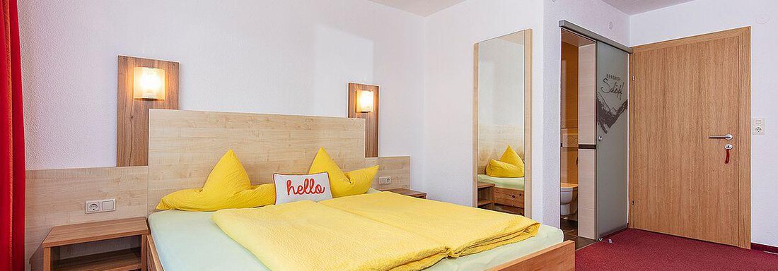 A cozy bedroom with a large bed and bright yellow blankets. The walls are painted in neutral colors, and there is a mirror as well as a wooden wardrobe. - Berghof Schöpf - Gries