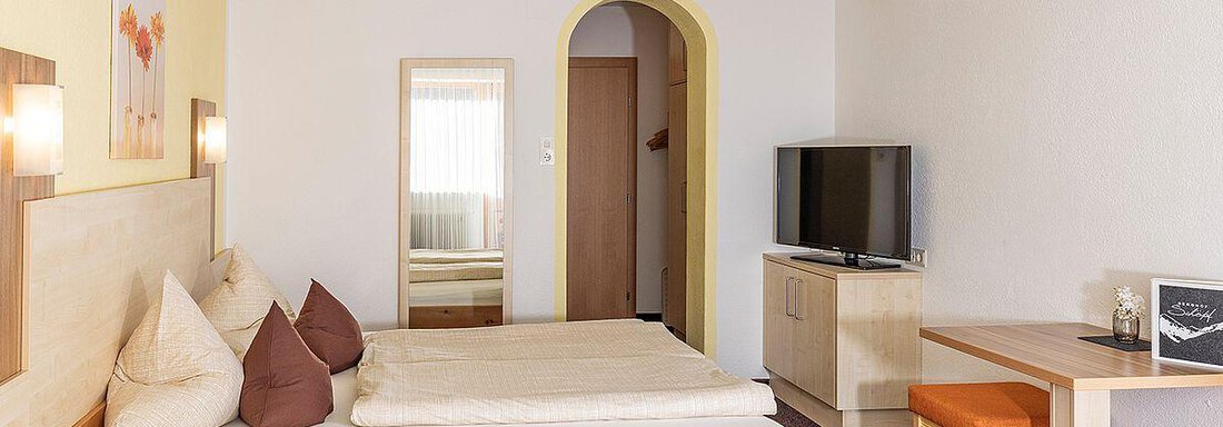 A cozy hotel room with wooden furniture and a double bed. There is a small table, a television, and a wall mirror. - Berghof Schöpf - Gries