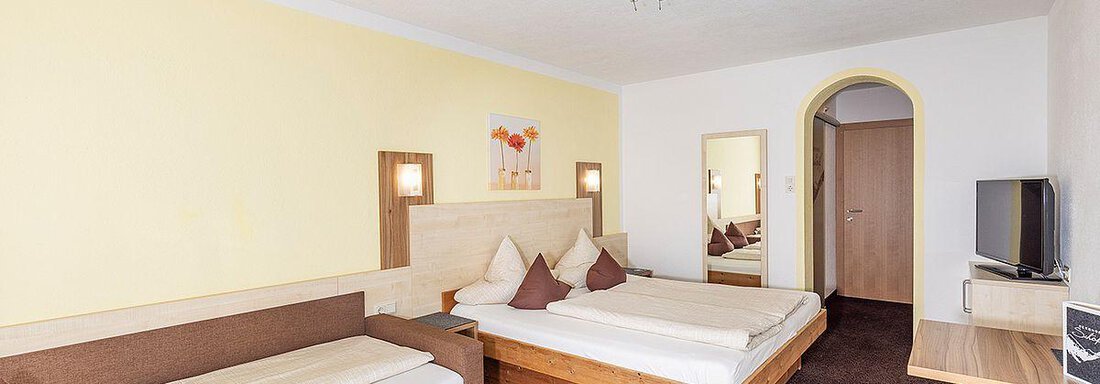 A bright hotel room with two beds and a cozy seating area. The walls are painted in soft colors, and there is a television as well as a mirror. - Berghof Schöpf - Gries