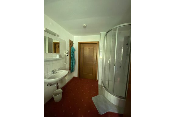 Ein helles Badezimmer mit einer Dusche, Waschbecken und einer Holzttür. Der Boden ist mit rotem Teppich ausgelegt und ein Handtuch hängt an einem Haken. - Haus Bergkristall - Gries