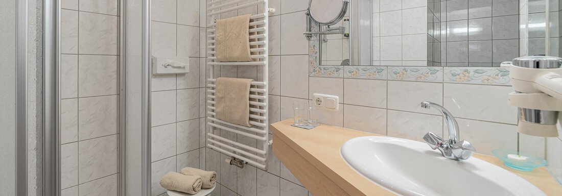 Ein modernes Badezimmer mit weißen Fliesen und einem großen Spiegel. Es verfügt über eine Dusche, ein Waschbecken und Handtücher auf einem Handtuchhalter. - Hotel Schöpf - Gries