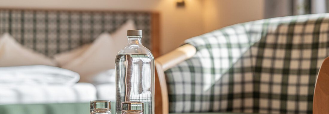 Ein gemütliches Zimmer mit einem Tisch, einer Glasflasche und zwei Gläsern. Im Hintergrund sind ein Bett und ein Sessel mit kariertem Muster zu sehen. - Hotel Schöpf - Gries