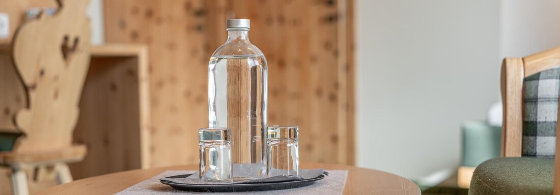 Ein Holzhaustisch mit einer Flasche Wasser und zwei Gläsern. Im Hintergrund sind Holzregale und eine grüne Couch zu sehen. - Hotel Schöpf - Gries