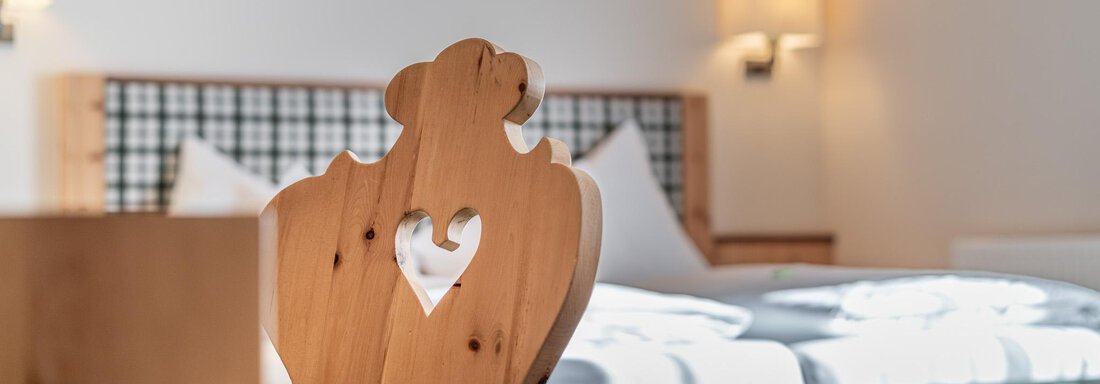 Ein gemütliches Schlafzimmer mit einem Holzstuhl, der ein Herzmotiv hat. Im Hintergrund ist ein großes Bett und sanftes Licht. - Hotel Schöpf - Gries