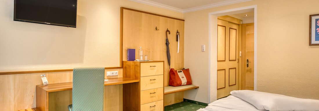Ein einfaches Zimmer mit einem Doppelbett, einem Schreibtisch und einem Fernseher. Die Wände sind hell und der Boden ist mit grünem Teppich ausgelegt. - Hotel Schöpf - Gries