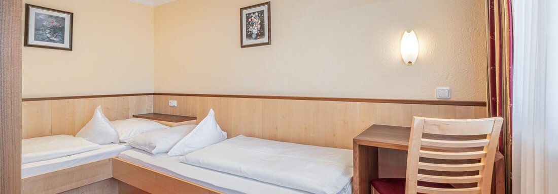 Ein einfaches Zimmer mit zwei Betten und einem Schreibtisch. Die Wände sind hell und die Teppichbodenfarbe ist rot. - Hotel Schöpf - Gries
