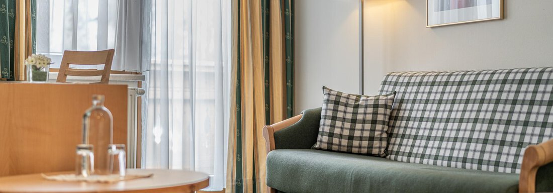 Ein gemütlicher Wohnraum mit einem grünen Sofa und einem Holztisch. Die Fenster sind mit Vorhängen verhängt und das Licht ist warm und einladend. - Hotel Schöpf - Gries