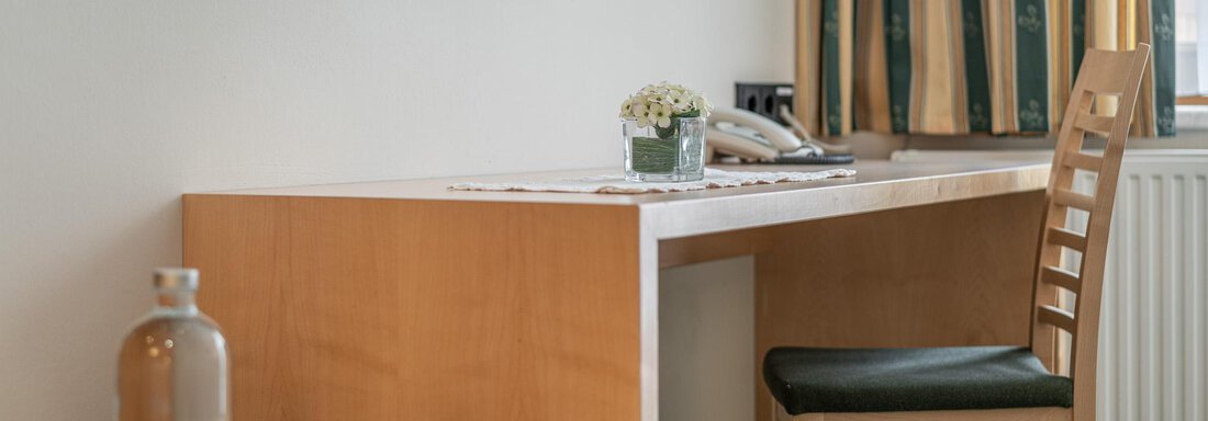 Ein bequemes Zimmer mit einem Holzschreibtisch und einem Stuhl. Auf dem Tisch steht eine kleine Pflanze und es gibt Gläser sowie eine Wasserflasche. - Hotel Schöpf - Gries