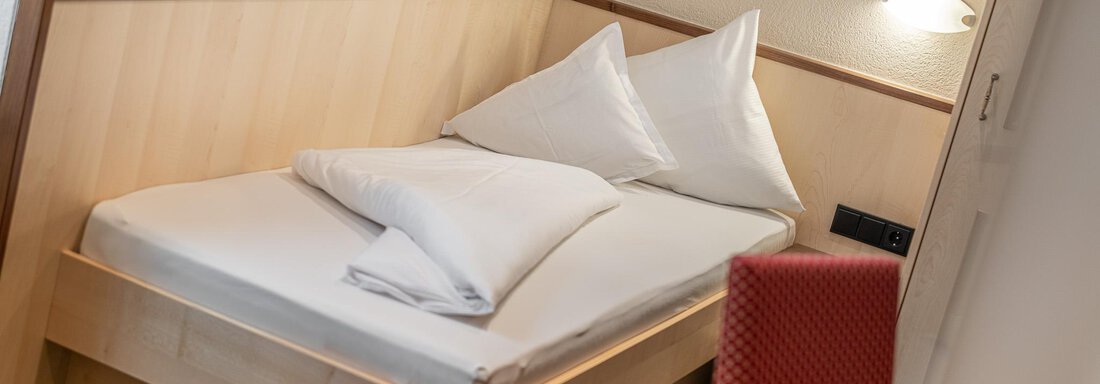 Ein einfaches Hotelzimmer mit einem Einzelbett und weißen Kissen. Die Wände sind hell und der Boden ist mit einem Teppich in grünem Muster ausgelegt. - Hotel Schöpf - Gries