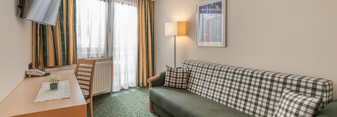 Ein gemütlicher Hotelzimmer mit einem Sofa, einem Tisch und einem Fernseher. Große Fenster lassen viel Tageslicht herein. - Hotel Schöpf - Gries