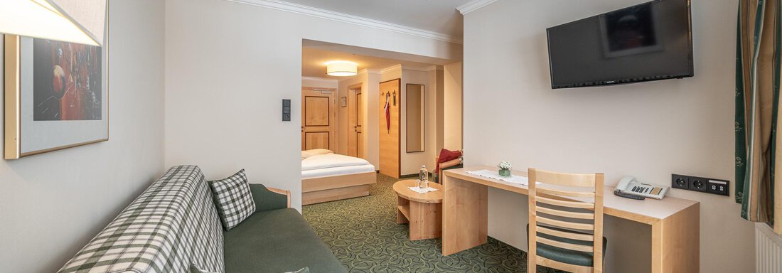 Ein gemütliches Zimmer mit einem Sofa, einem Tisch und einem Fernseher. Im Hintergrund sieht man ein Schlafzimmer mit einem Doppelbett. - Hotel Schöpf - Gries