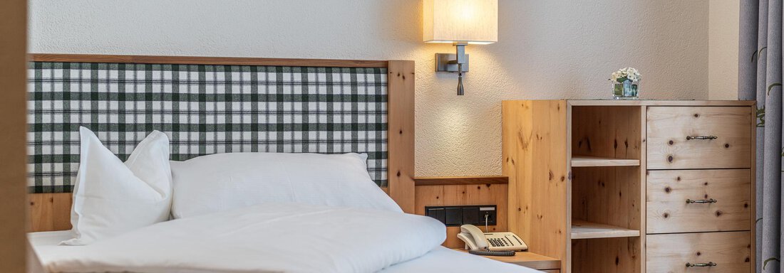 Ein gemütliches Hotelzimmer mit einem großen Bett und Holzverkleidung. Neben dem Bett steht eine Kommode und ein Nachttisch mit einer Lampe. - Hotel Schöpf - Gries