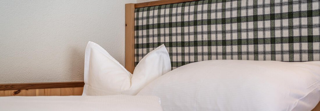 Ein gemütliches Bett mit weißen Kissen und einer karierten Kopfstütze. Die Bettwäsche ist frisch und ordentlich. - Hotel Schöpf - Gries