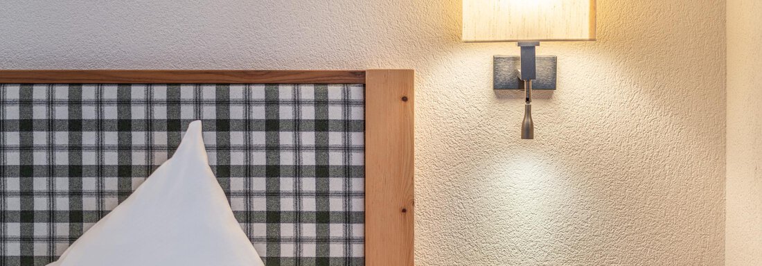 Ein gemütliches Schlafzimmer mit einem Holzkopfteil und einem Kissen. An der Wand hängt eine moderne Lampe, die sanft beleuchtet. - Hotel Schöpf - Gries