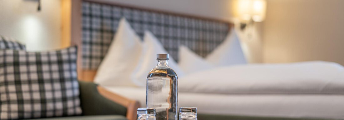 Ein gemütliches Hotelzimmer mit einem Tisch, auf dem eine Wasserflasche und Gläser stehen. Im Hintergrund sind ein Bett und dekorative Kissen sichtbar. - Hotel Schöpf - Gries