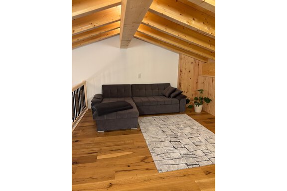A cozy living room with a gray couch and a rug on wooden floor. The ceiling is made of wood with visible beams. - KLAMMERHOF - Gries