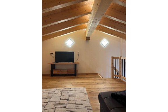 A stylish attic with wooden beams and large windows. A television is on a modern TV table in a cozy atmosphere. - KLAMMERHOF - Gries