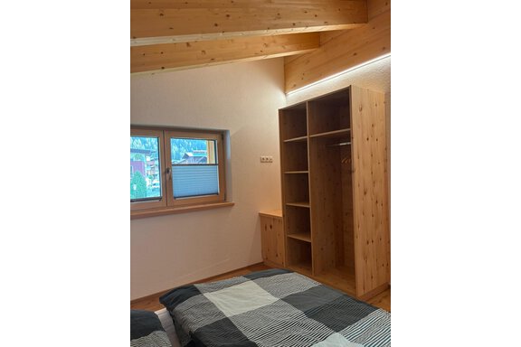 A modern bedroom with wooden walls and a large window. 
There is a bed with a checked bedspread and a wooden wardrobe. - KLAMMERHOF - Gries