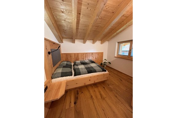 A cozy bedroom with a wooden bed and checkered bedding. The walls and ceiling are made of wood, and there is a window with a nice view. - KLAMMERHOF - Gries