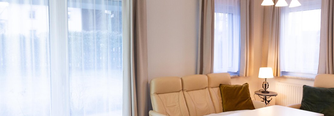 Ein gemütliches Wohnzimmer mit hellen Vorhängen und einem bequemen Sofa. Ein Tisch und eine Lampe vervollständigen die freundliche Atmosphäre. - Ferienhaus Neurauter - Haiming