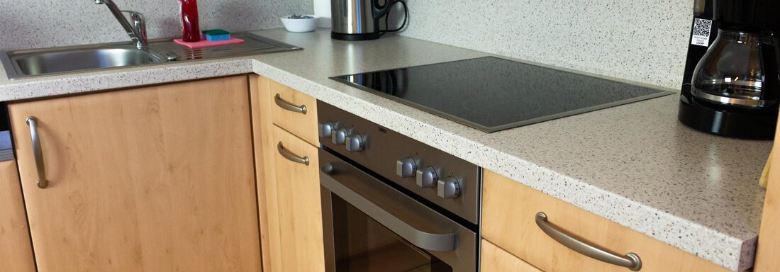 Eine moderne Küche mit Holzschränken und einer hellen Arbeitsfläche. Zu sehen sind ein Herd, ein Wasserkocher und eine Kaffeemaschine. - Ferienhaus Neurauter - Haiming