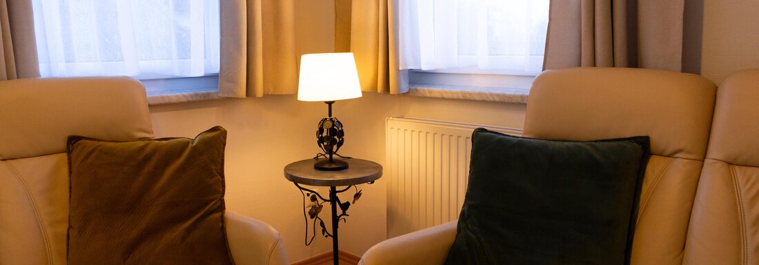 Ein gemütlicher Raum mit beigen Sesseln und einem kleinen Tisch. Eine Lampe sorgt für warmes Licht neben den Fenstern. - Ferienhaus Neurauter - Haiming