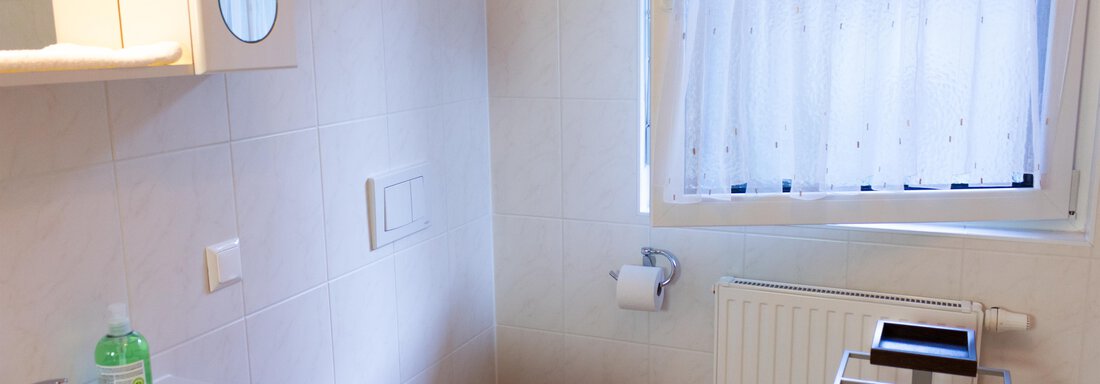 Ein helles Badezimmer mit Dusche, Waschbecken und WC. Es hat ein Fenster mit einem lichtdurchlässigen Vorhang und einen Handtuchhalter. - Ferienhaus Neurauter - Haiming
