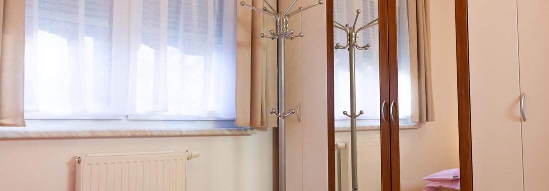 Ein helles Schlafzimmer mit einem Bett und einem großen Kleiderschrank. Der Raum hat Fenster mit Vorhängen und einen Garderobenständer. - Ferienhaus Neurauter - Haiming
